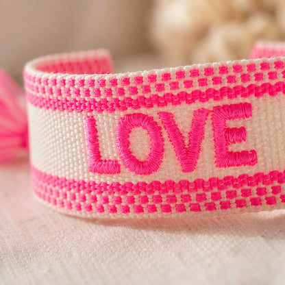 Nahaufnahme des bestickten LOVE-Schriftzugs auf cremefarbenem gewebtem Armband, Detailansicht der neonpinken Stickerei und Streifenmuster

