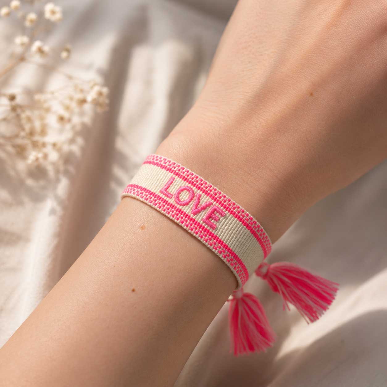 Cremefarbenes LOVE Armband mit neonpinken Streifen und rosa Quasten am Handgelenk getragen, verstellbares Webarmband mit Schleierkraut im Hintergrund
