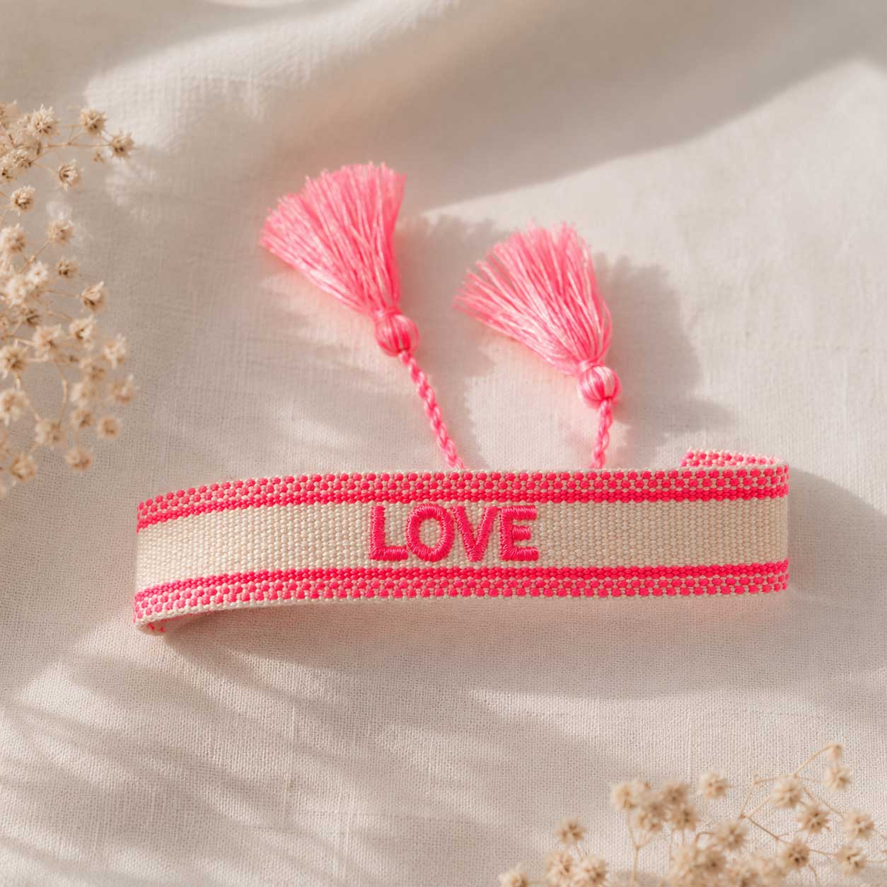 Cremefarbenes gewebtes LOVE Armband mit neonpinken Streifen und zwei rosa Quasten auf weißem Leinenstoff im Sonnenlicht, verstellbares Festival-Armband
