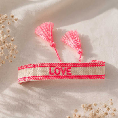 Cremefarbenes gewebtes LOVE Armband mit neonpinken Streifen und zwei rosa Quasten auf weißem Leinenstoff im Sonnenlicht, verstellbares Festival-Armband
