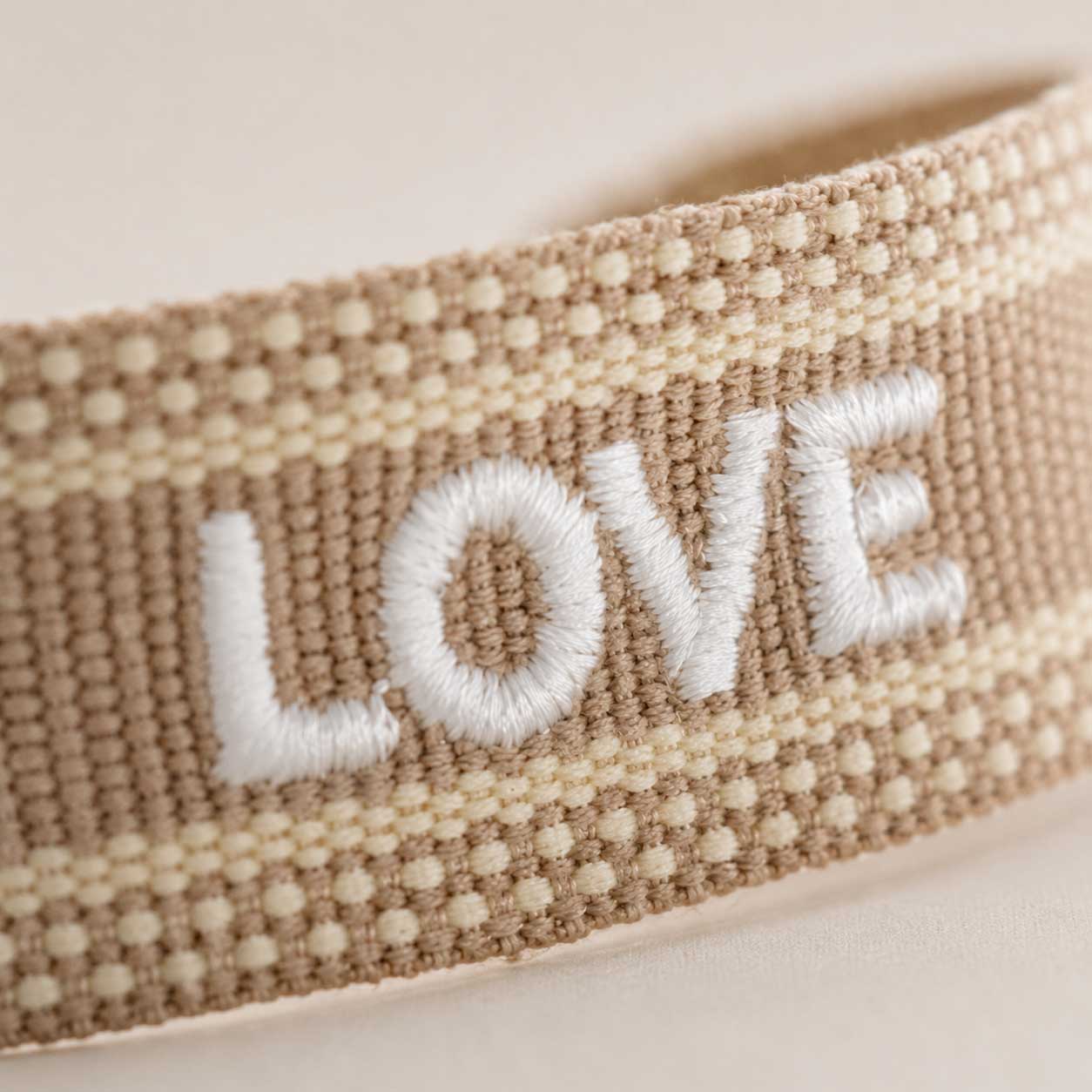 Nahaufnahme gestickter Schriftzug „Love" in Weiß auf beigem Webarmband – Saltyblush Modeschmuck

