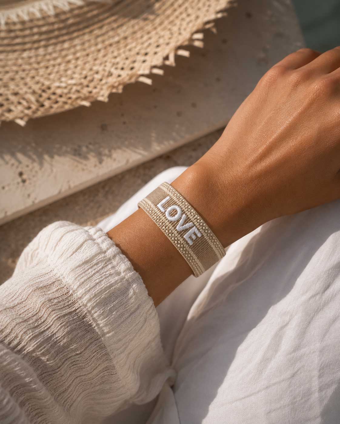 Beiges Love Statement Armband am Handgelenk mit Strohhut im Hintergrund – Sommer Schmuck Damen, Saltyblush
