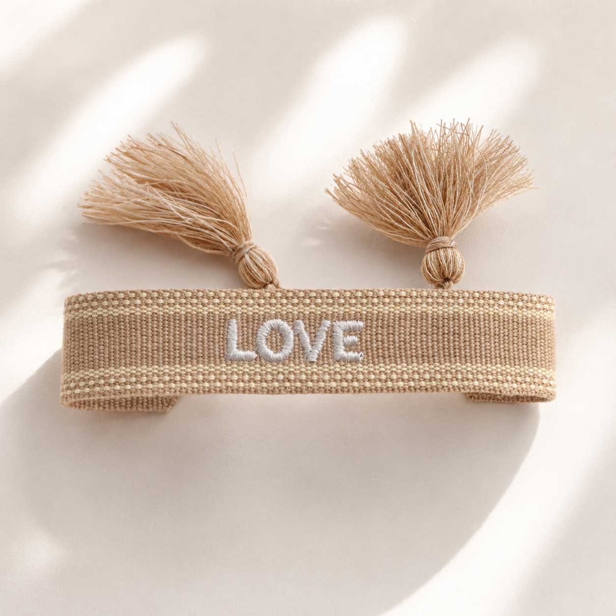 Love Armband aufgeklappt mit zwei beigen Quasten und weißem Schriftzug – Flatlay Produktfoto, Saltyblush
