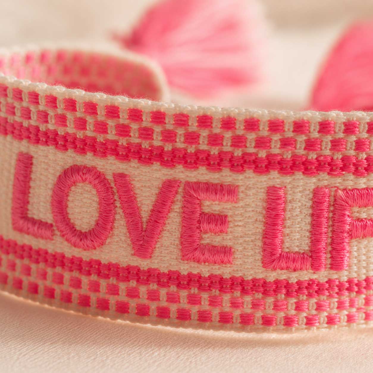 Nahaufnahme gestickter Schriftzug „Love Life" in Neonpink auf cremefarbenem Webarmband – Saltyblush Modeschmuck
