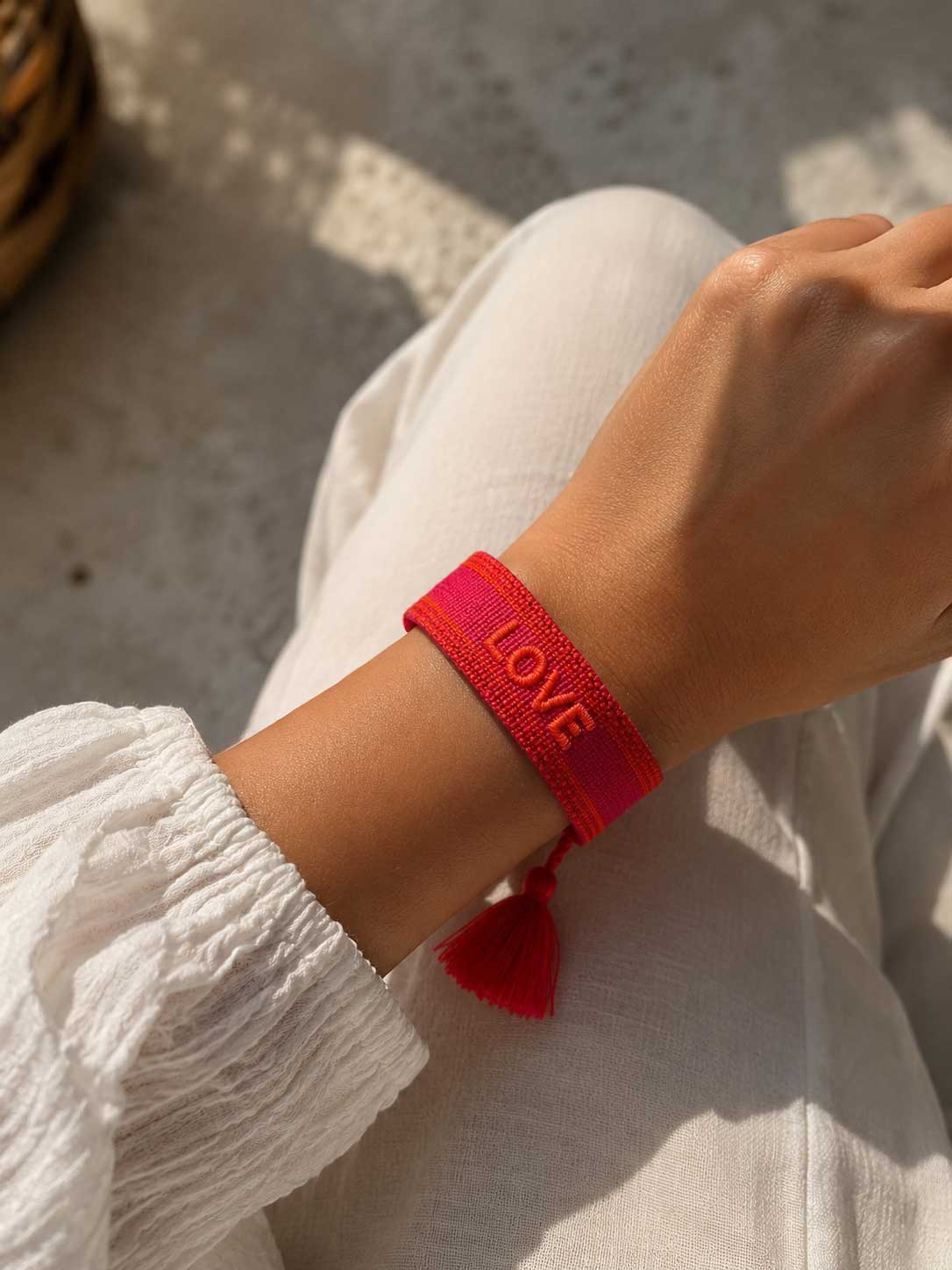 Rotes gewebtes LOVE Armband am Handgelenk, getragen zu weißem Sommeroutfit im Freien
