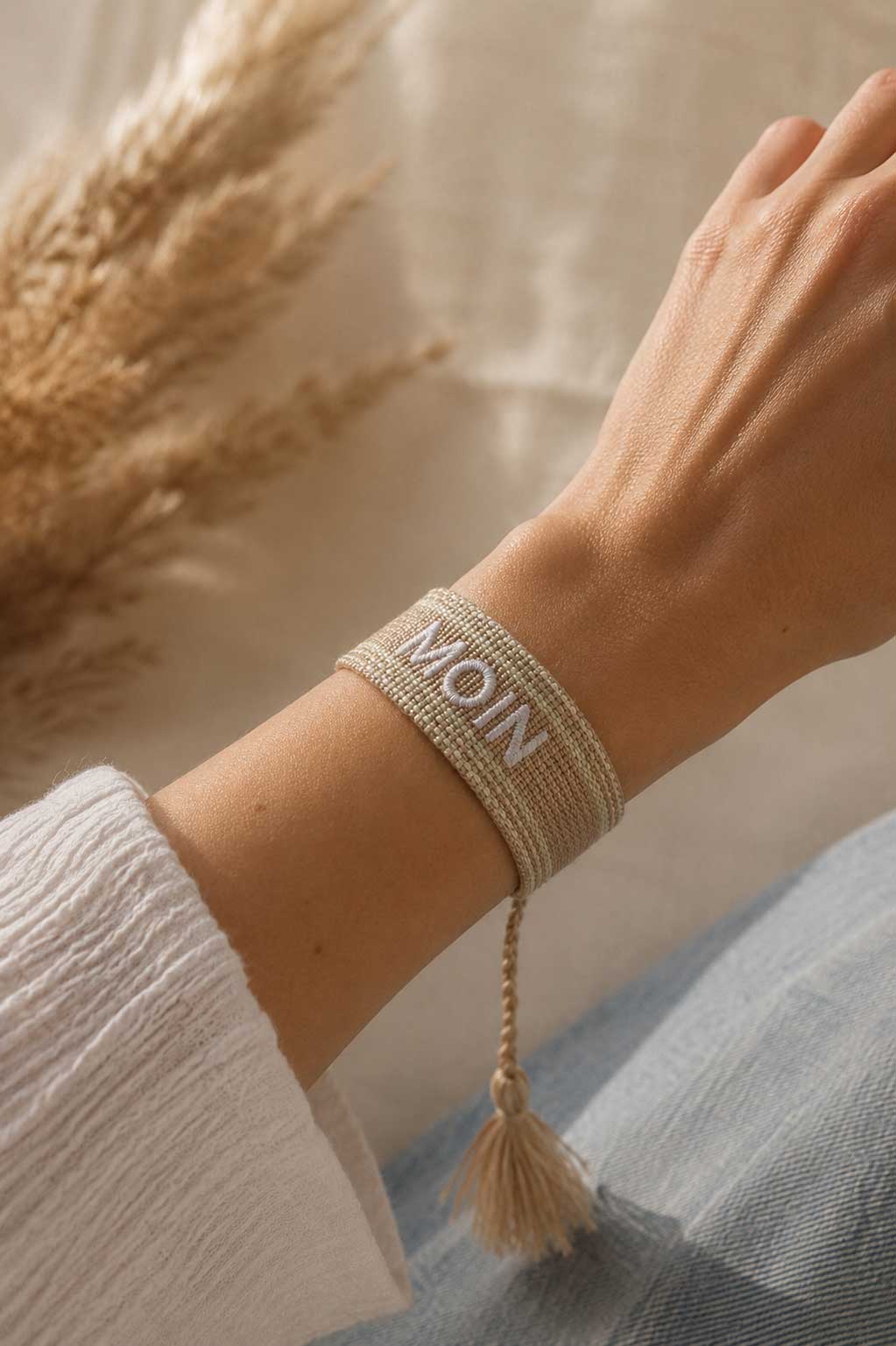 Beiges MOIN Armband mit cremefarbener Quaste am Handgelenk getragen, weißes Oberteil, Pampasgras im Hintergrund – Saltyblush
