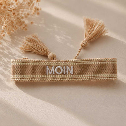 Beiges MOIN Armband mit zwei cremefarbenen Quasten aufgeklappt auf hellem Stoff, Trockenblumen im Hintergrund – Saltyblush
