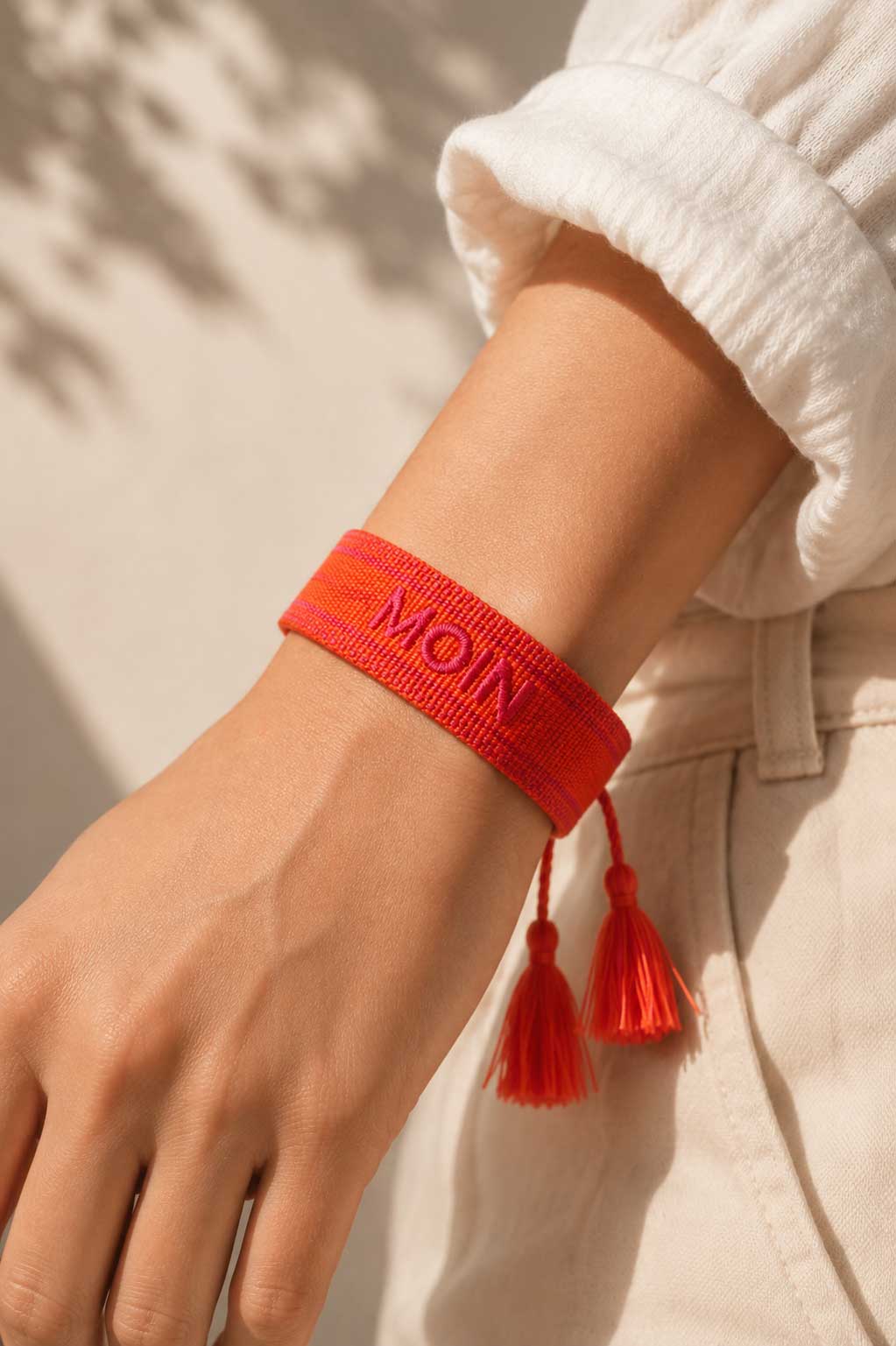 Oranges MOIN Armband mit zwei roten Quasten am Handgelenk getragen, weißes Leinenoberteil, sandiger Hintergrund – Saltyblush
