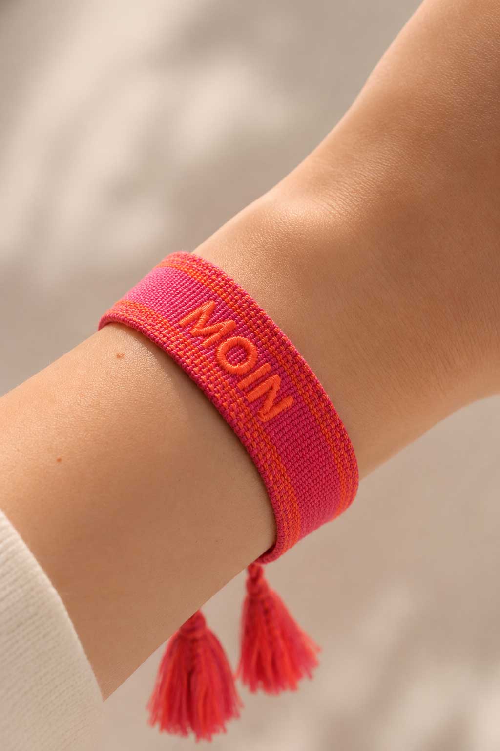 Pinkes MOIN Armband mit roten Quasten am Handgelenk getragen, weißes Oberteil, heller Hintergrund – Saltyblush
