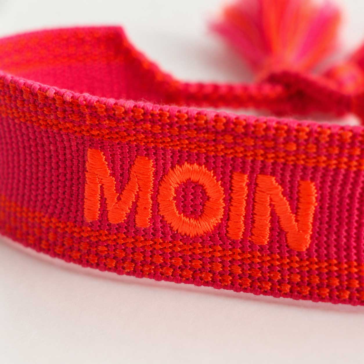 Nahaufnahme der orangen MOIN Stickerei auf pink-rotem gewebtem Textilarmband – Saltyblush
