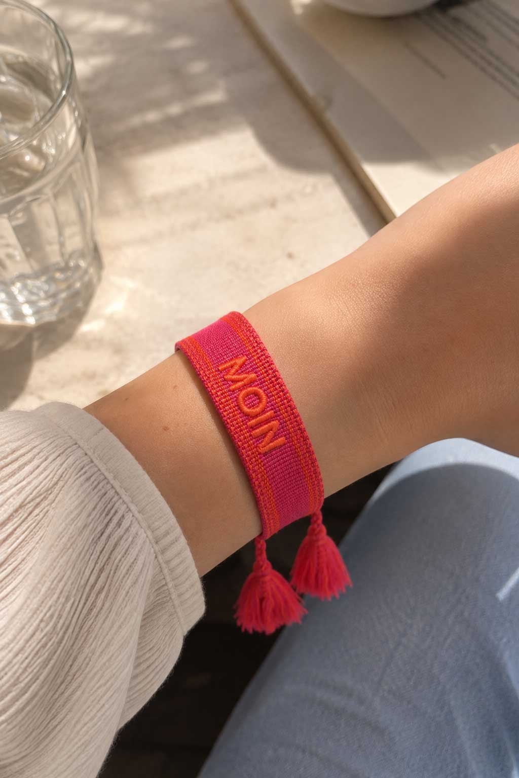 Pinkes MOIN Armband mit roten Quasten am Handgelenk getragen, weißes Oberteil und Jeans, Sonnenlicht auf Holzuntergrund – Saltyblush
