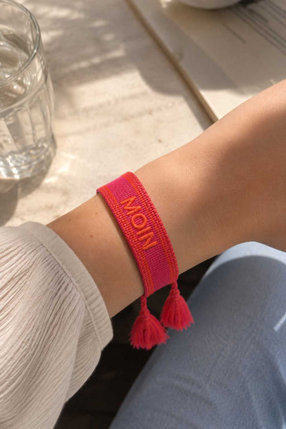 Pinkes MOIN Armband mit roten Quasten am Handgelenk getragen, weißes Oberteil und Jeans, Sonnenlicht auf Holzuntergrund – Saltyblush
