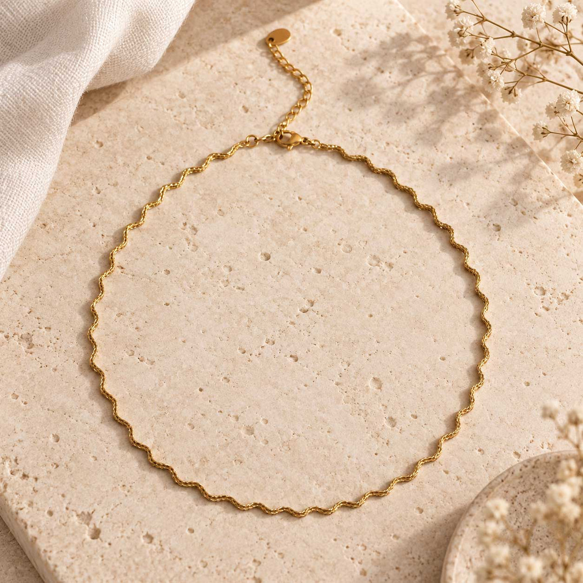 Ocean Line goldene Halskette – wellenförmige Wellenkette aus vergoldetem Edelstahl mit Karabinerverschluss und Verlängerungskette, auf Travertin-Stein mit Schleierkraut und Leinentuch, Produktfoto
