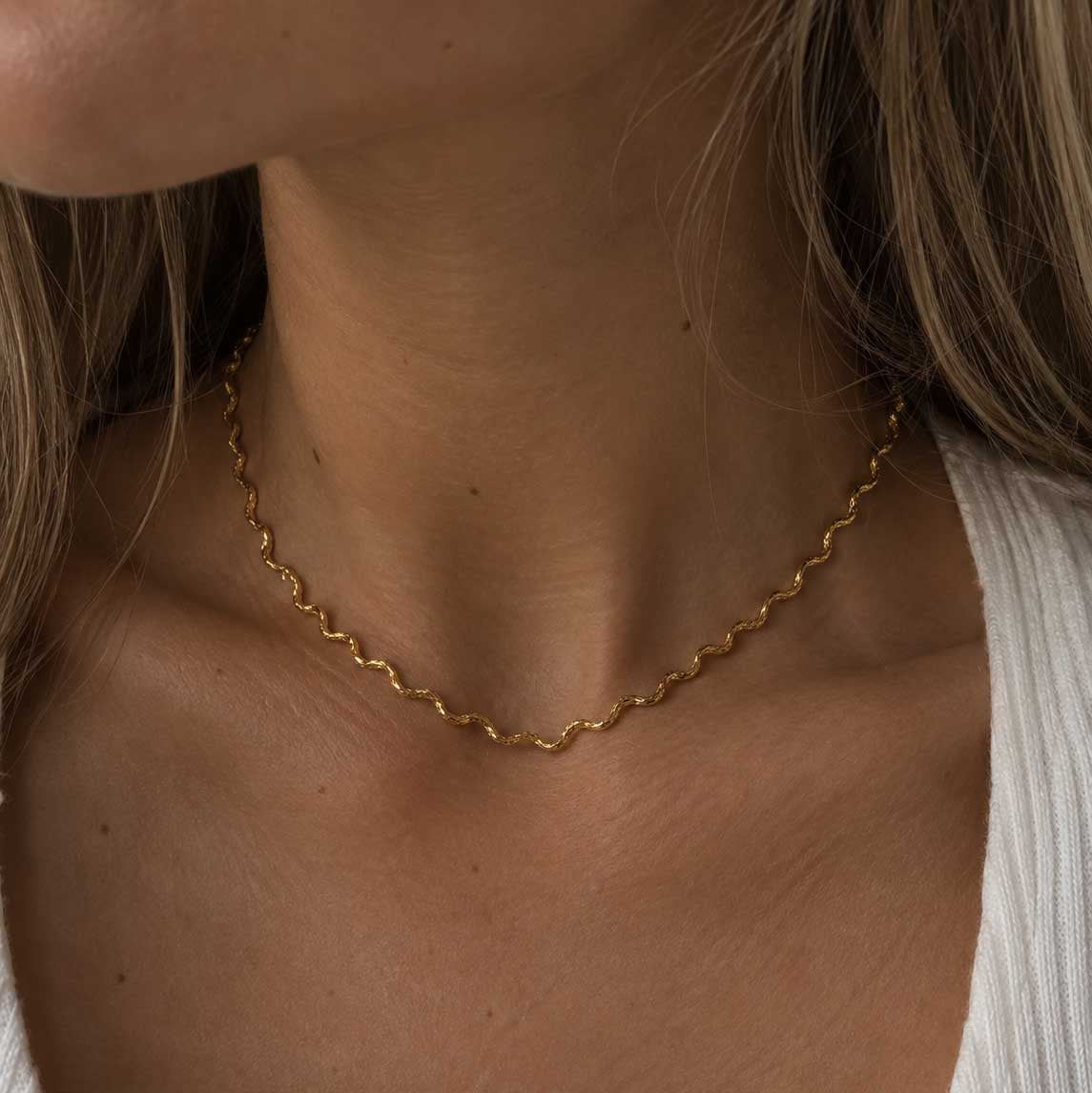 Ocean Line goldene Halskette getragen – blonde Frau, Nahaufnahme Hals, zarte wellenförmige Goldkette aus Edelstahl, weißes Rippenstrick-Oberteil, cremefarbener Hintergrund
[AI]