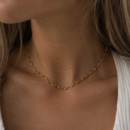 Ocean Line goldene Halskette getragen – blonde Frau, Nahaufnahme Hals, zarte wellenförmige Goldkette aus Edelstahl, weißes Rippenstrick-Oberteil, cremefarbener Hintergrund
[AI]