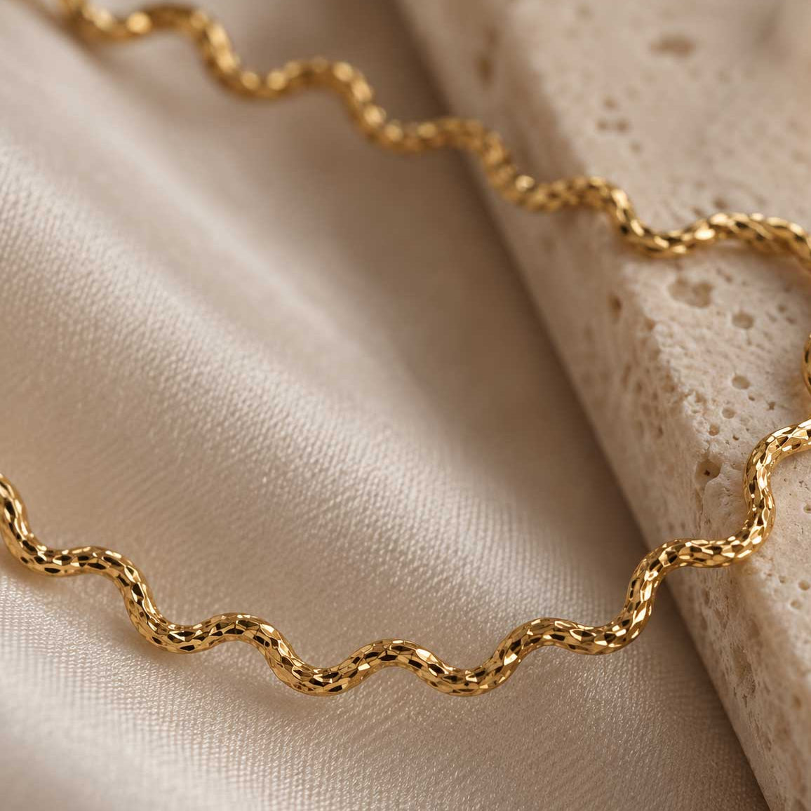 Ocean Line goldene Halskette Detailaufnahme – facettierte wellenförmige Glieder aus vergoldetem Edelstahl mit feinen Lichtreflexen, auf cremefarbenem Satin und Travertin, Makroansicht
