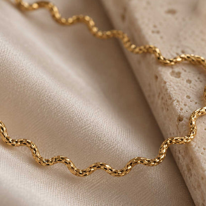 Ocean Line goldene Halskette Detailaufnahme – facettierte wellenförmige Glieder aus vergoldetem Edelstahl mit feinen Lichtreflexen, auf cremefarbenem Satin und Travertin, Makroansicht

