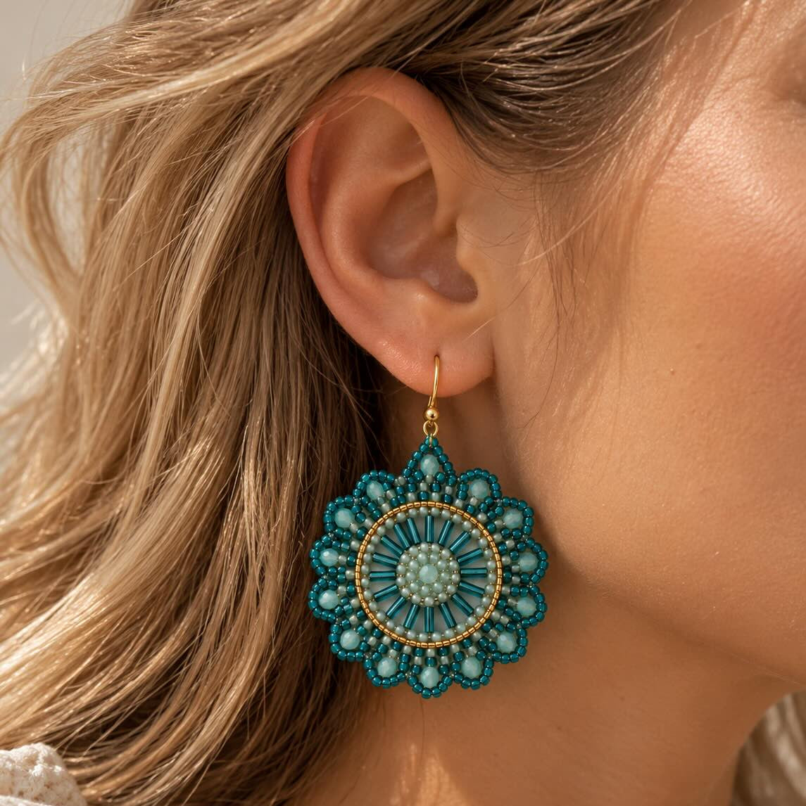 Ocean Mandala goldene Perlenohrringe getragen – blonde Frau, Nahaufnahme Ohr, großes rundes türkis-weißes Mandala-Perlenohrring mit goldenem Einfassungsring und Hakenverschluss, weißer Strickpullover, heller Innenraum
[AI]