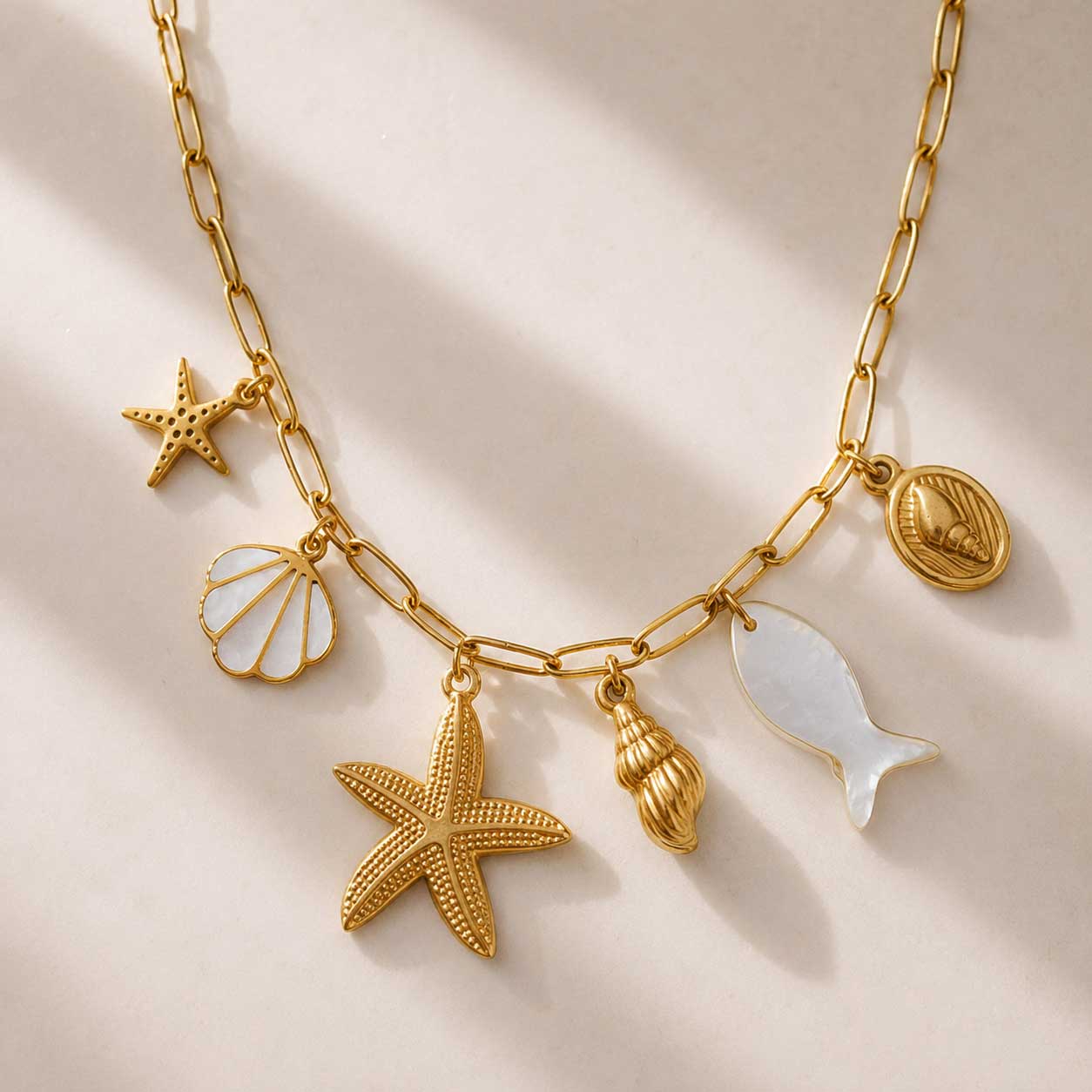 Goldene Ocean Treasures Charm-Halskette auf cremefarbenem Seidenstoff im Sonnenlicht – Seestern, Perlmutt-Muschel, Schnecke, Perlmutt-Fisch und ovaler Muschel-Anhänger auf vergoldeter Paperclip-Kette
