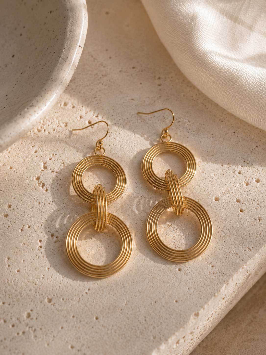 Goldene Doppelring-Ohrhänger mit geripptem Drahtmuster auf cremefarbenem Stein mit weißem Seidenstoff im Sonnenlicht, vergoldete Edelstahl-Ohrringe mit zwei verbundenen Kreisen
