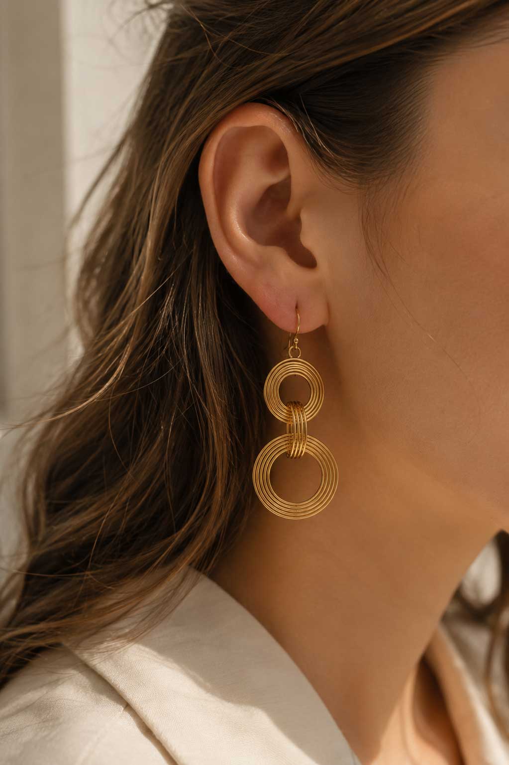 Goldene Doppelring-Ohrhänger am Ohr getragen, vergoldete Edelstahl-Ohrringe mit zwei gerippten Kreisen und goldenem Ohrhaken zu braunem Haar und weißem Outfit
