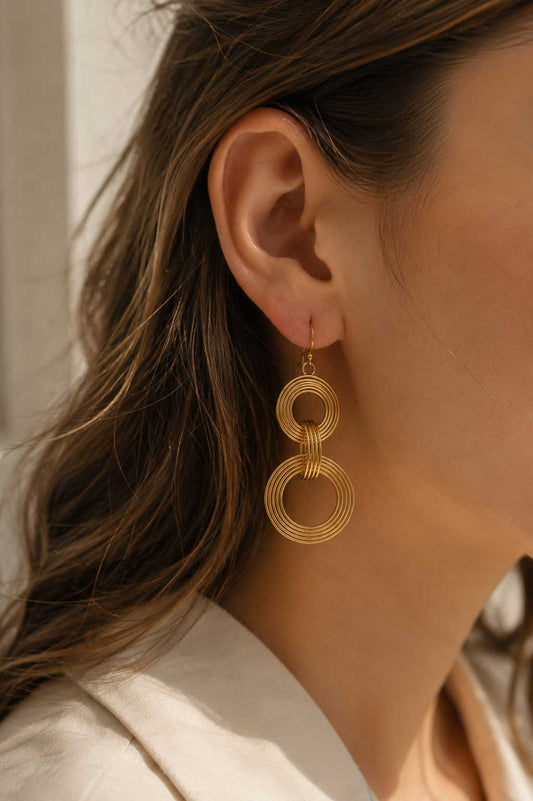 Goldene Doppelring-Ohrhänger am Ohr getragen, vergoldete Edelstahl-Ohrringe mit zwei gerippten Kreisen und goldenem Ohrhaken zu braunem Haar und weißem Outfit
