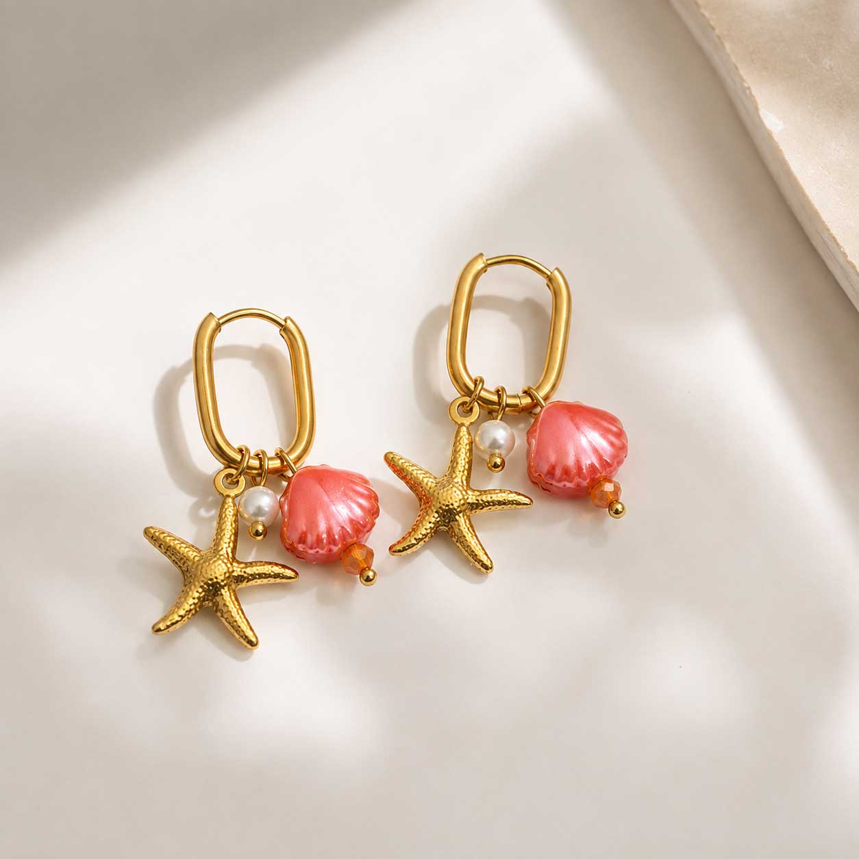 Pearl and Sea goldene Ohrringe mit Seestern, pinker Muschel und Perle auf cremefarbenem Hintergrund
