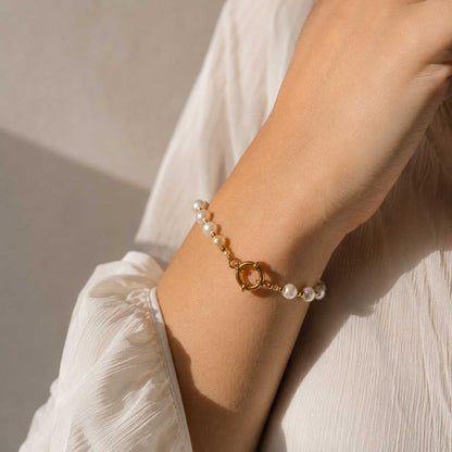 Goldenes Pearl Glow Perlenarmband am Handgelenk getragen, weißes Süßwasserperlen-Armband mit vergoldetem Edelstahl-Ringverschluss zu weißem Leinenoberteil im Sonnenlicht

