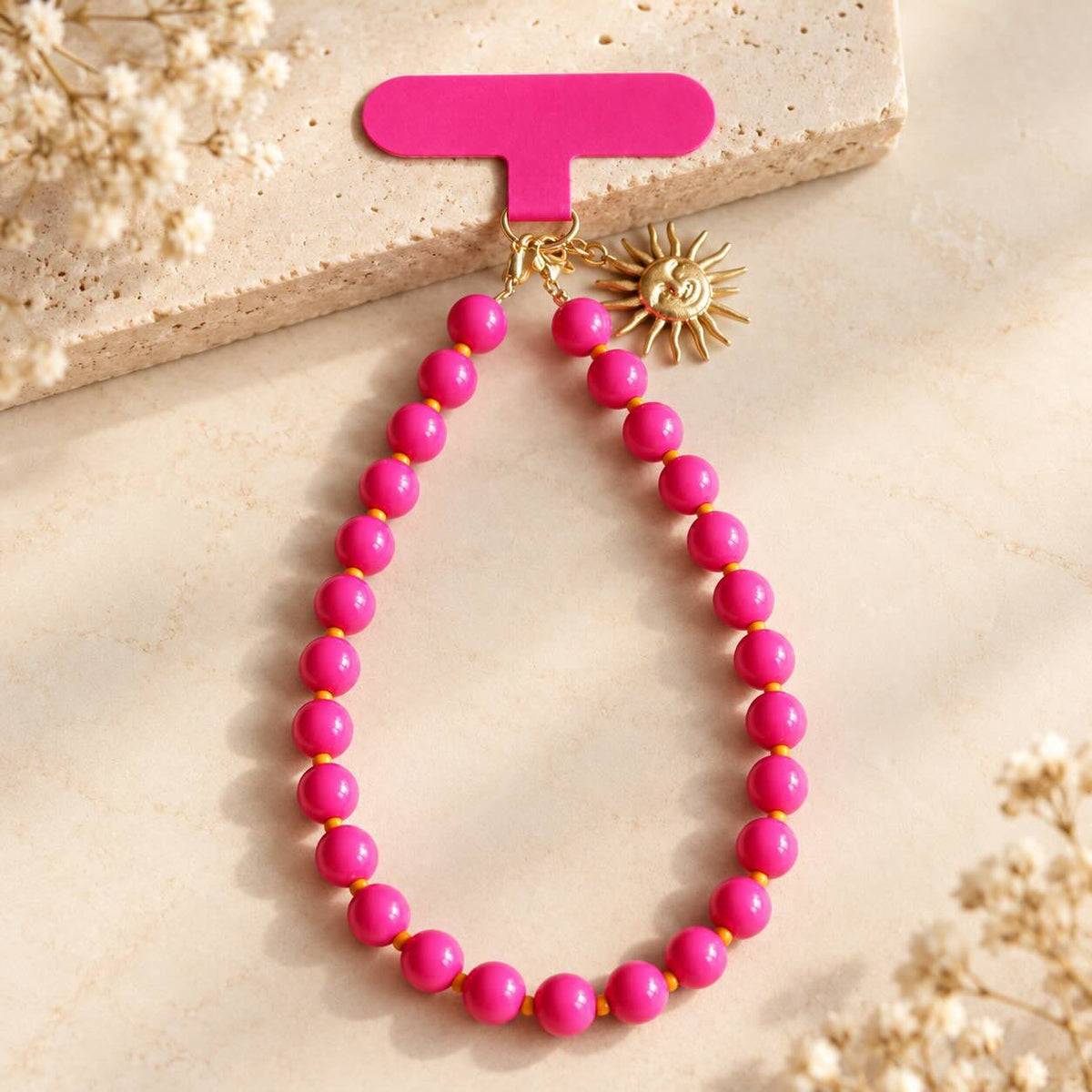 Pinkfarbener Phone Charm aus runden Perlen mit orangefarbenen Zwischenperlen und goldenem Sonnenanhänger, auf hellem Stein arrangiert.
