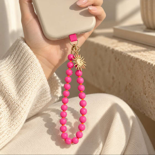 Pinkfarbener Perlen-Phone-Charm mit goldenem Sonnenanhänger hängt an einer hellen Handyhülle, die in der Hand gehalten wird.
