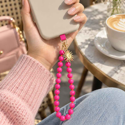 Pinkfarbener Phone Charm mit goldenem Sonnenanhänger an einer hellen Handyhülle, in der Hand gehalten neben einer Tasse Kaffee.
