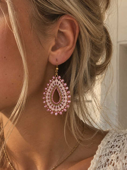 Rosewater goldene Perlenohrringe getragen – blonde Frau, Nahaufnahme Ohr, rosa Boho-Tropfenohrringe mit goldenem Hakenverschluss, heller Hintergrund

