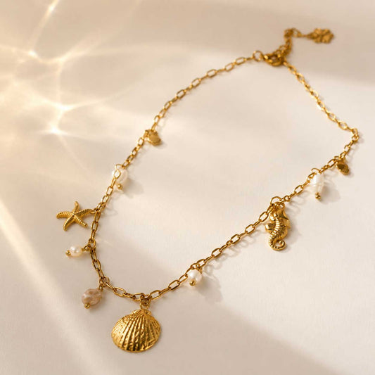 Goldene maritime Halskette – Salt Breeze Halskette von Saltyblush, Edelstahlkette mit Muschel-, Seestern-, Seepferdchen- und Perlenanhängern, Flatlay auf weißem Stoff im Sonnenlicht
