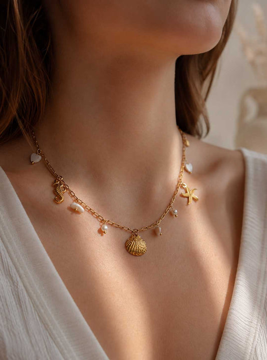 Salt Breeze Halskette getragen – goldene maritime Charm-Kette von Saltyblush am Hals einer Frau mit braunem Haar, Muschel- und Seesternanhänger zu weißem Oberteil
