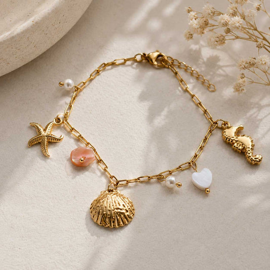 Goldenes Charm-Armband mit Muschel, Seestern, Seepferdchen, rosa Perlmutt-Stein, weißem Herzanhänger und Süßwasserperlen auf cremefarbenem Stein mit Schleierkraut
