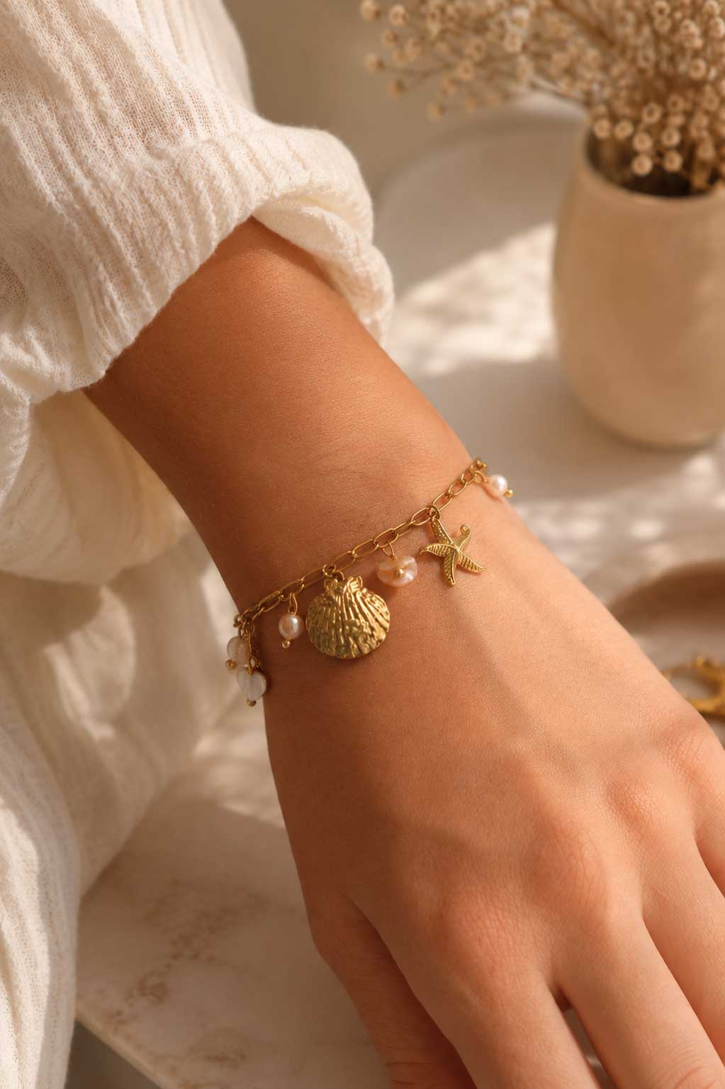 Goldenes Meer-Charm-Armband am Handgelenk getragen mit Muschel, Seestern und Süßwasserperlen zu weißem Rüschenoberteil, vergoldete Paperclip-Kette mit Karabinerverschluss
