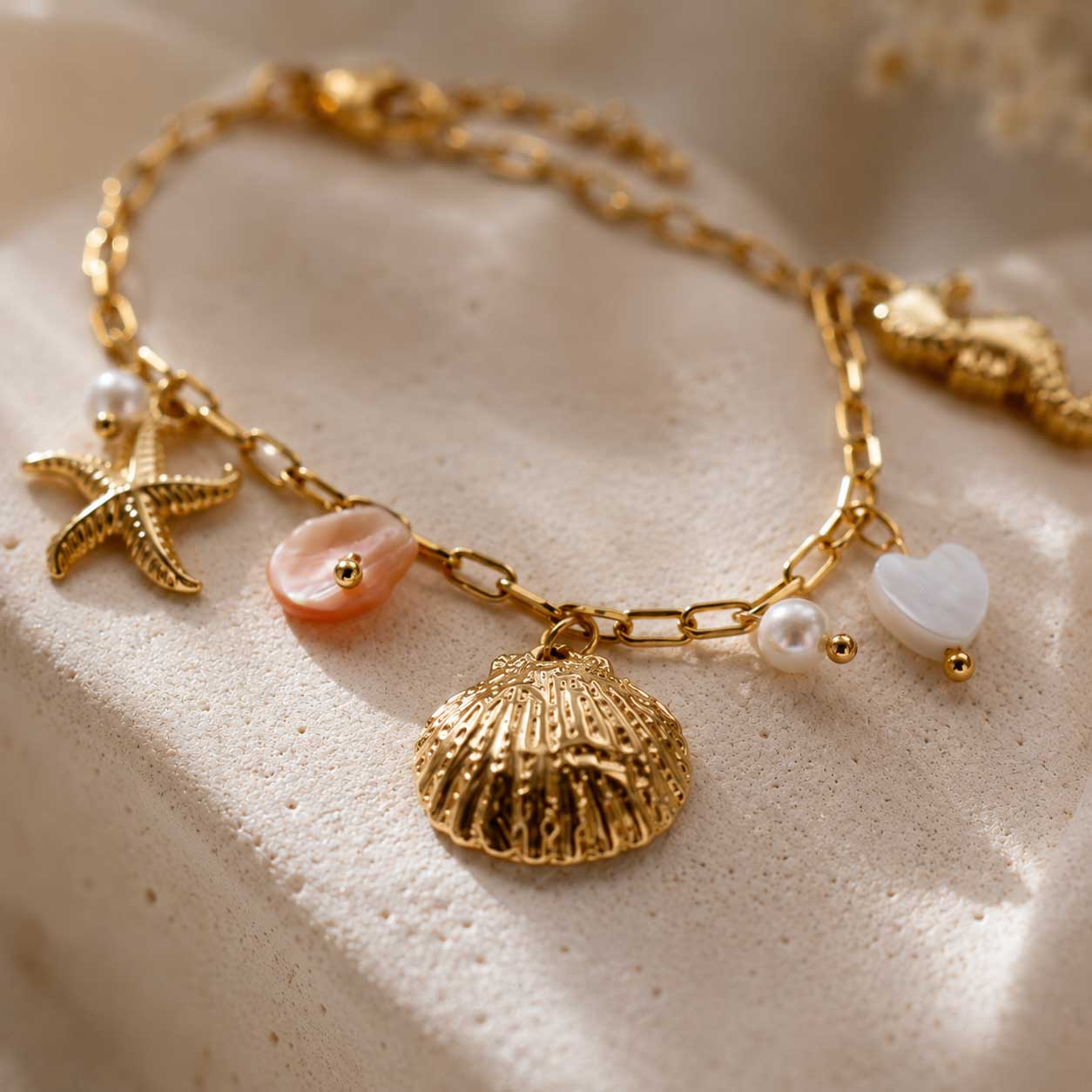 Nahaufnahme goldenes Charm-Armband auf cremefarbenem Stein – Detailansicht Muschel, Seestern, Seepferdchen, rosa Perlmutt-Stein, weißes Herz und Süßwasserperlen auf vergoldeter Paperclip-Kette
