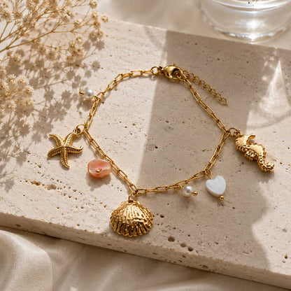 Goldenes Meer-Charm-Armband auf Travertin-Stein im Sonnenlicht mit Schleierkraut – Muschel, Seestern, Seepferdchen, rosa Perlmutt, weißes Herz und Perlen auf vergoldeter Paperclip-Kette
