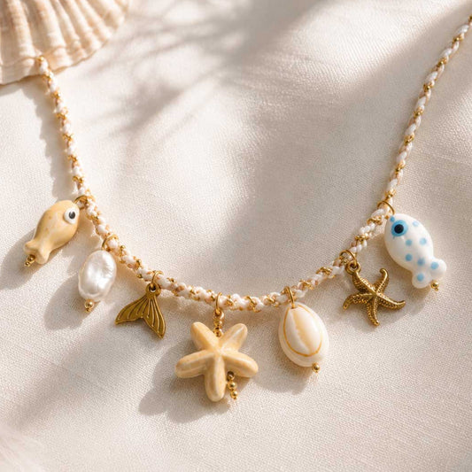 Salty Star Halskette Flatlay – maritime Charm-Kette von Saltyblush mit Seestern, Muschel, Meerjungfrauenflosse, Perle und Fisch-Charms auf weißem Stoff mit Palmenschatten
