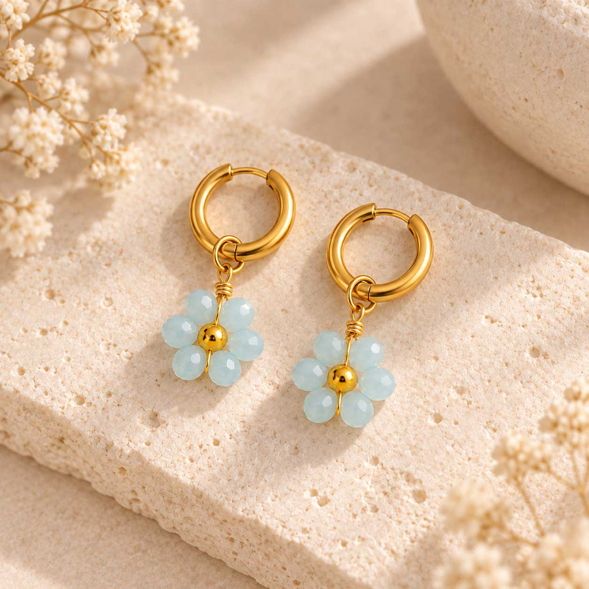 Sea Blossom goldene Ohrringe – hellblaue Blütenanhänger aus facettierten Perlen an goldener Creole, auf Travertin-Stein mit Trockenblumen
