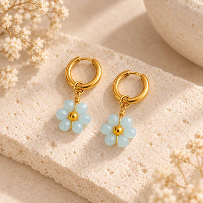 Sea Blossom goldene Ohrringe – hellblaue Blütenanhänger aus facettierten Perlen an goldener Creole, auf Travertin-Stein mit Trockenblumen
