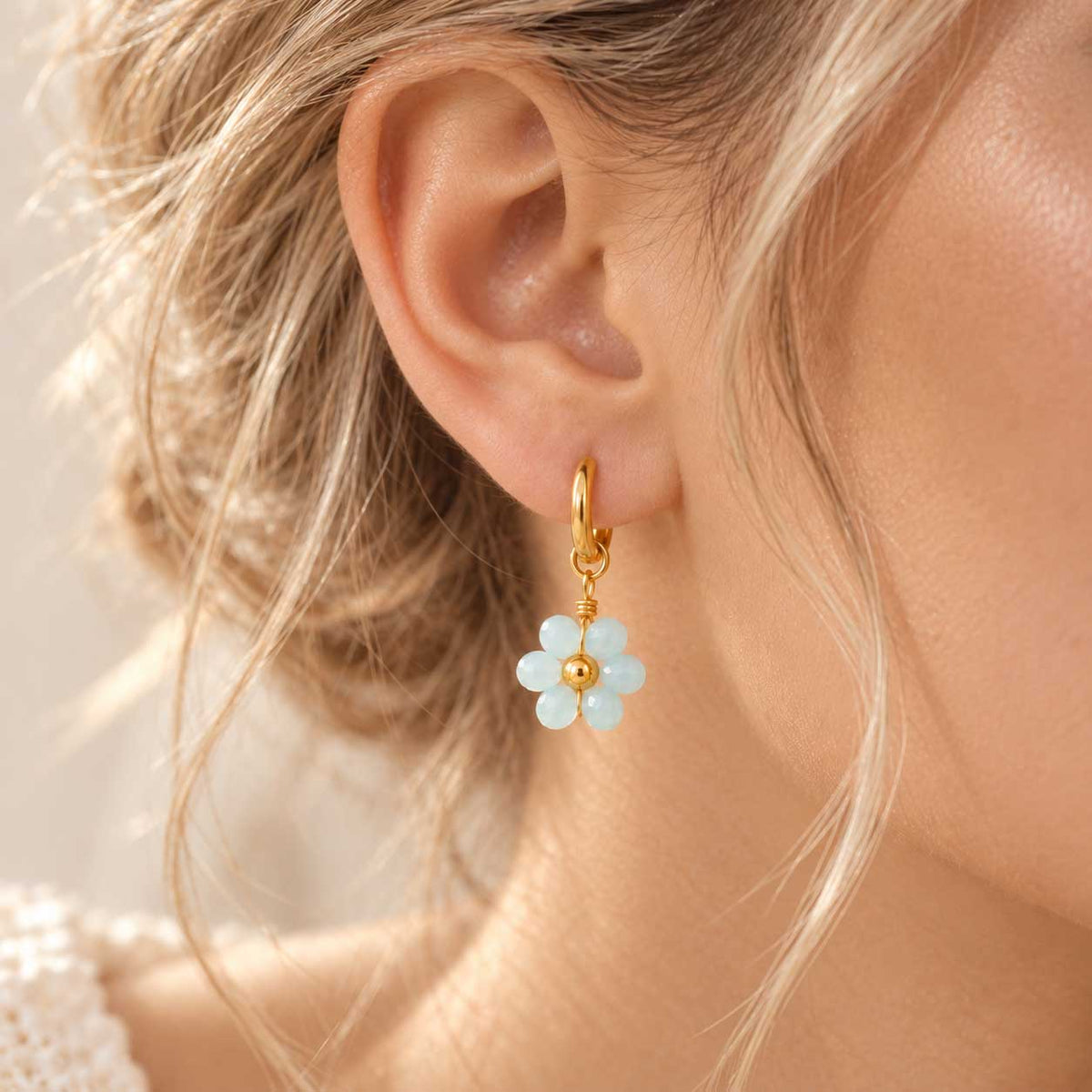 Sea Blossom goldene Ohrringe getragen – blonde Frau, Nahaufnahme Ohr, hellblauer Blütenanhänger an goldener Edelstahl-Creole
[AI]