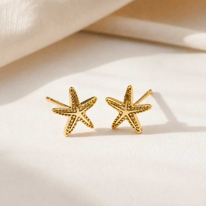 Goldene Seestern Ohrringe – Sea Spirit Stecker von Saltyblush, strukturierte Seestern-Stecker aus Edelstahl mit Kugeldetails, Produktfoto auf weißem Stoff
