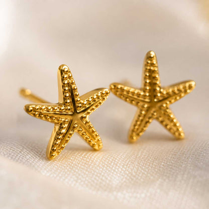 Nahaufnahme goldene Seestern Stecker – Sea Spirit Ohrringe von Saltyblush, detaillierte Oberflächenstruktur mit Kugelrand, Detailansicht auf weißem Stoff
