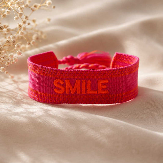 Pinkes Statement Armband mit oranger SMILE Stickerei auf cremefarbenem Stoff, Trockenblumen im Hintergrund – Saltyblush
