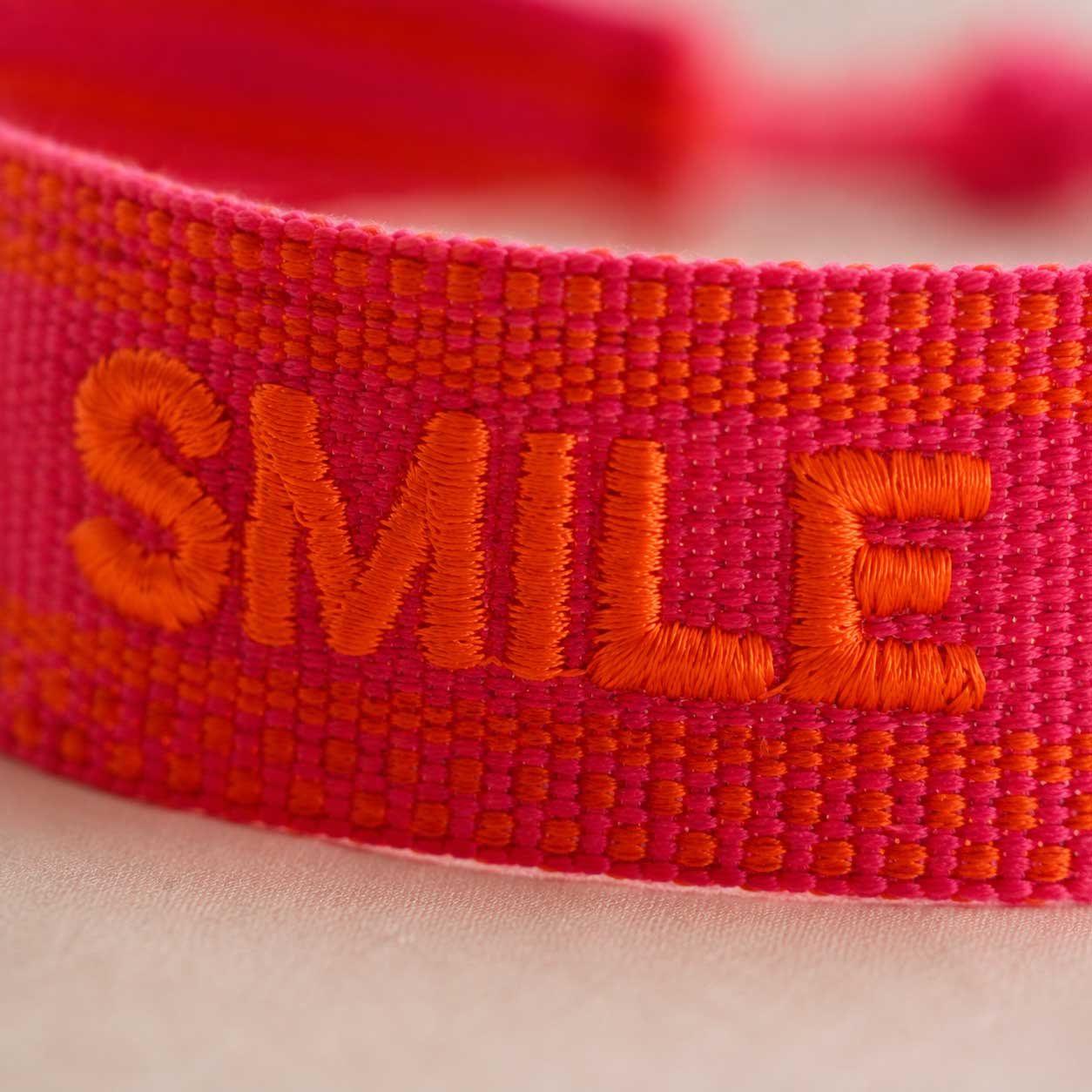 Nahaufnahme der orangen SMILE Stickerei auf pinkfarbenem Textilarmband – Saltyblush
