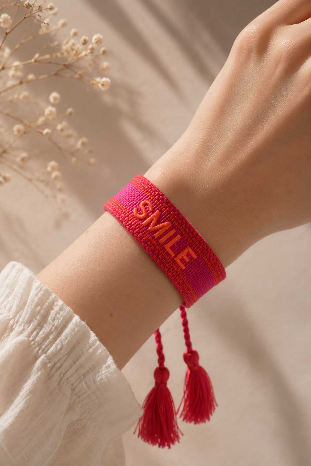 Pinkes SMILE Armband mit roten Quasten am Handgelenk getragen, weißes Kleid, Trockenblumen im Hintergrund – Saltyblush
