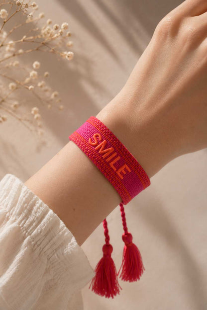 Pinkes SMILE Armband mit roten Quasten am Handgelenk getragen, weißes Kleid, Trockenblumen im Hintergrund – Saltyblush
