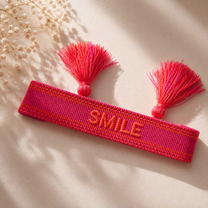 Pinkes SMILE Statement Armband mit zwei pinken Quasten aufgeklappt auf hellem Stoff, Draufsicht – Saltyblush
