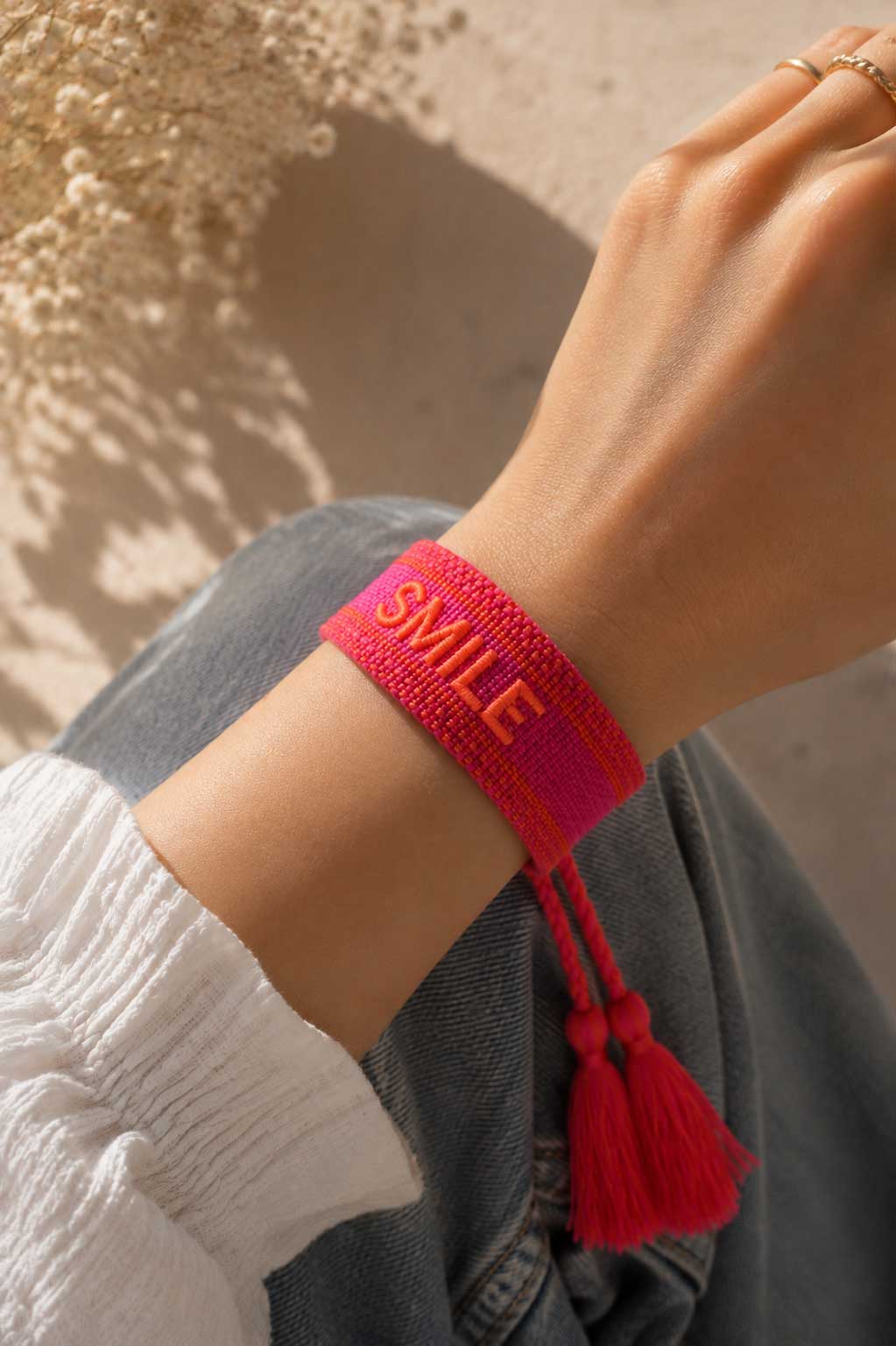 Pinkes SMILE Armband mit roten Quasten am Handgelenk getragen, weißes Oberteil und Jeans, Sonnenlicht – Saltyblush
